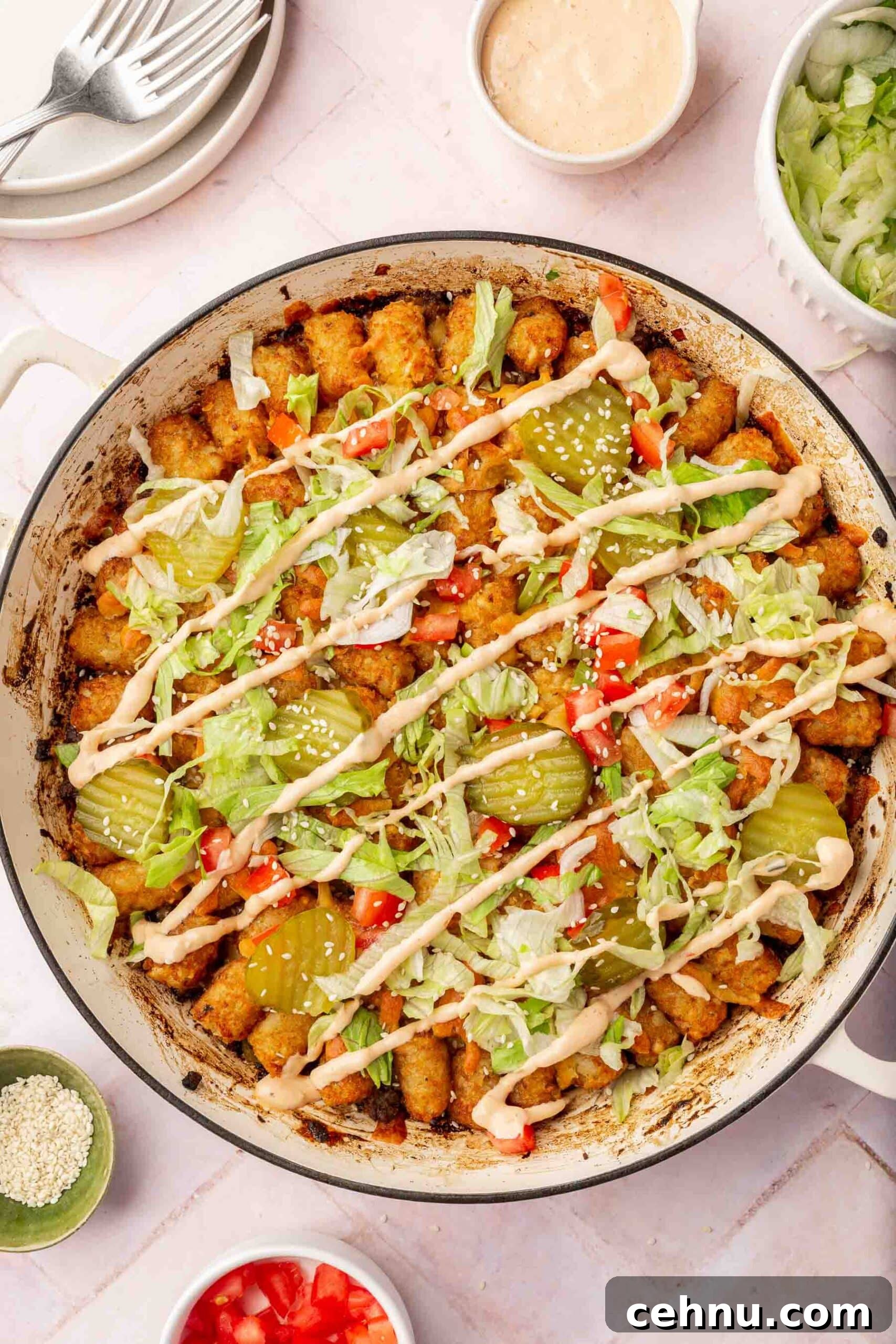 Big mac casserole in a white braising pan topped with lettuce, tomatoes, pickles, big mac sauce and sesame seeds.