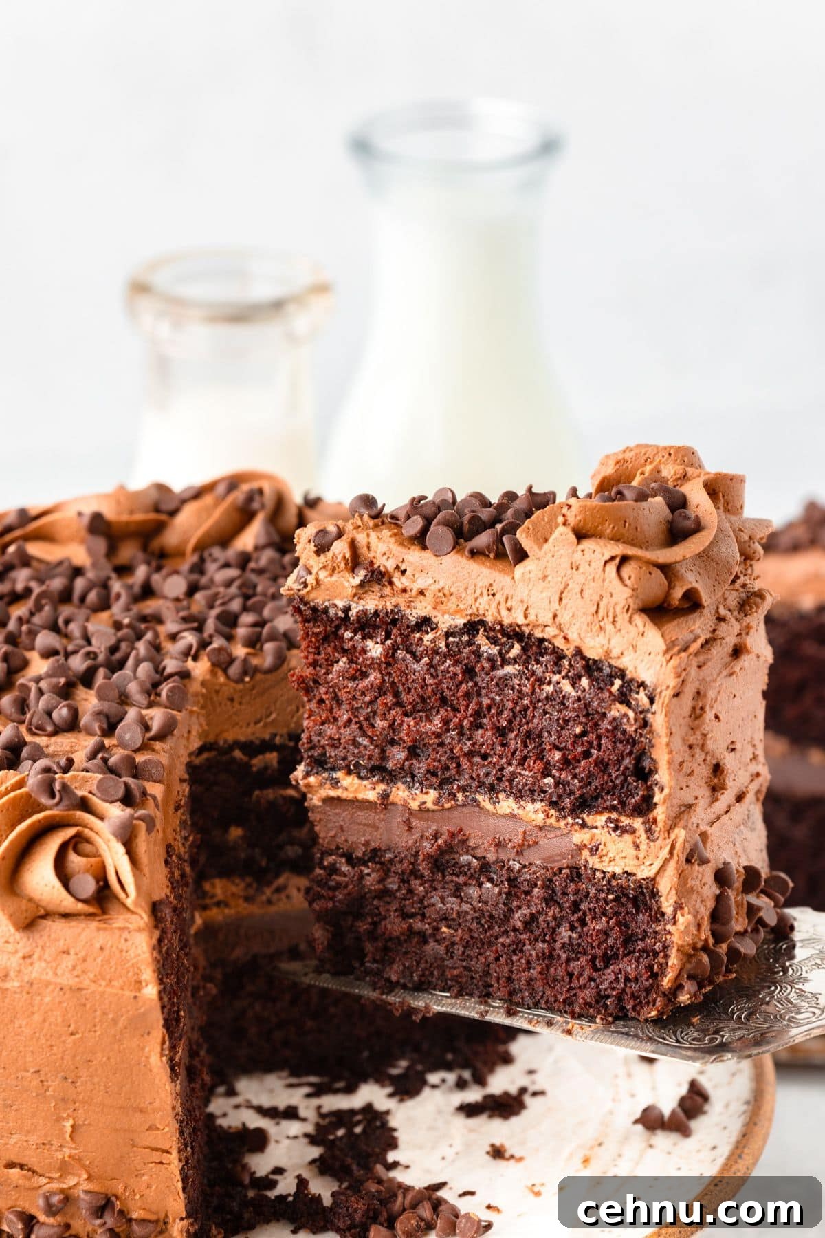 Decadent Triple Chocolate Cake Recipe 2 A rich chocolate cake with a slice being lifted out, topped with chocolate chips and swirls of chocolate frosting. Two layers are visible, with creamy chocolate filling in between. Blurred background shows bottles of milk.
