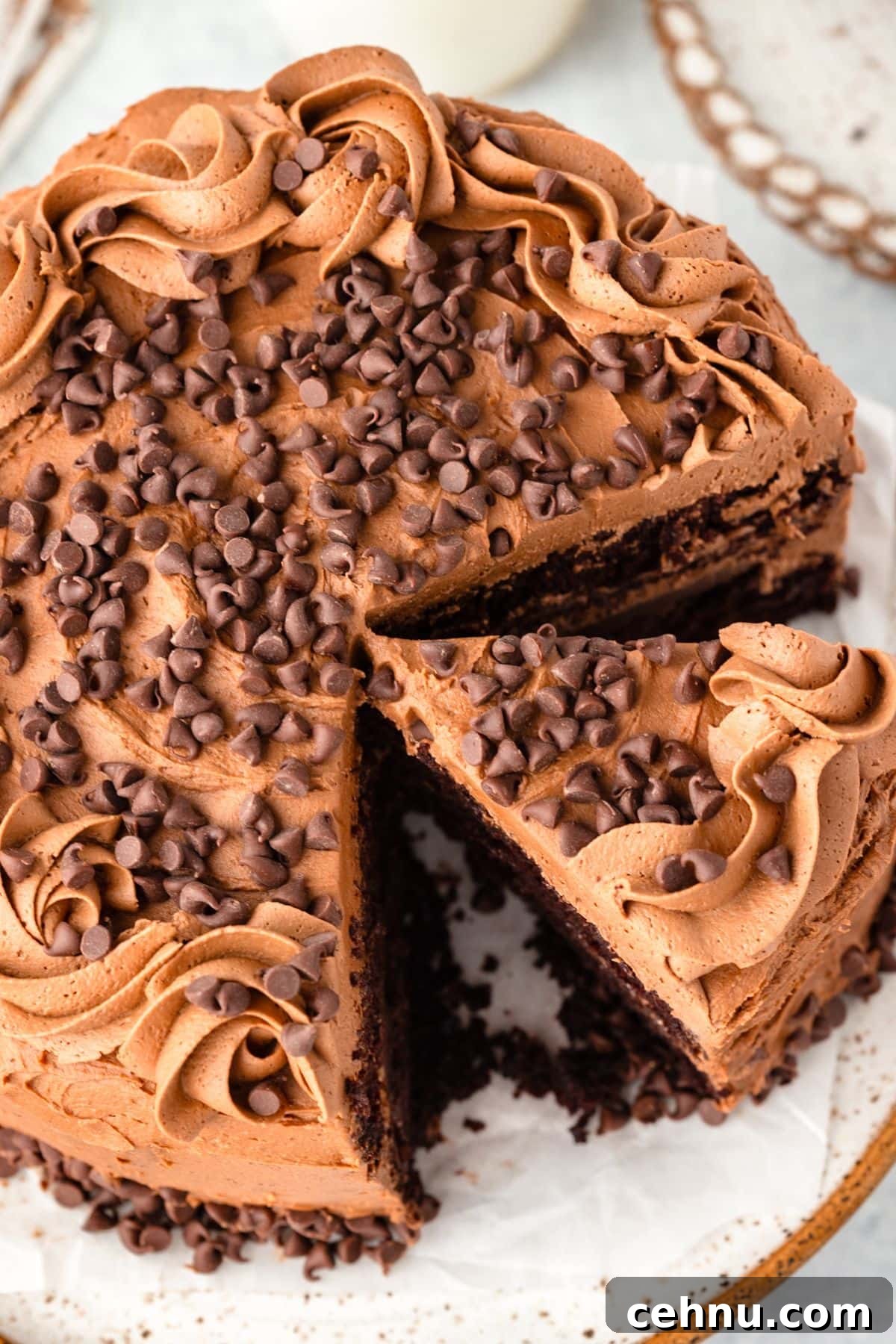Decadent Triple Chocolate Cake Recipe 3 A chocolate cake with a slice cut out, topped with chocolate frosting and chocolate chips. Swirls of frosting decorate the edges, and the cake sits on a parchment-lined plate.