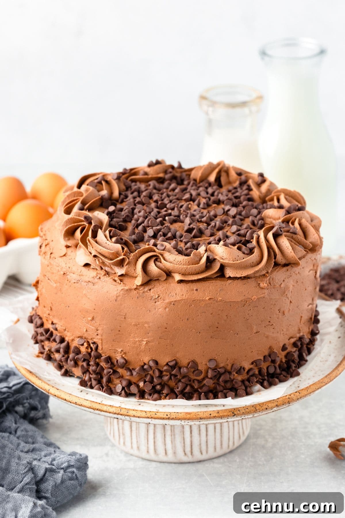 Decadent Triple Chocolate Cake Recipe 8 A round chocolate cake with chocolate frosting is topped with chocolate chips. It sits on a striped ceramic cake stand. In the background, there are fresh eggs, a bottle of milk, and a glass carafe. A textured grey cloth is nearby.