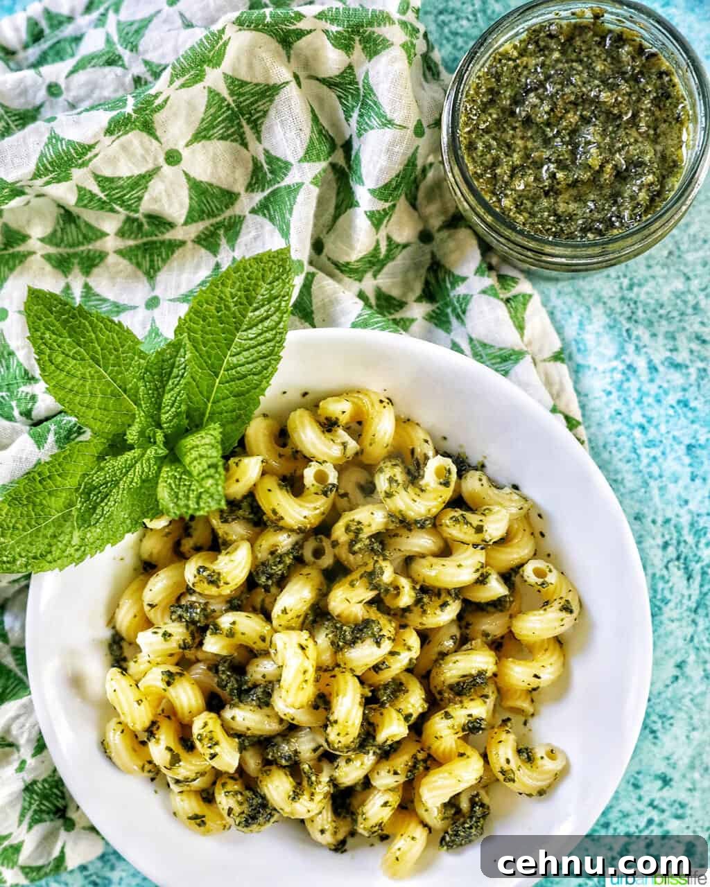 white bowl with pasta and mint pesto with mint leaves as garnish, with a jar of mint pesto in the top right corner, all on a bright blue background with green and white napkin to the left side.