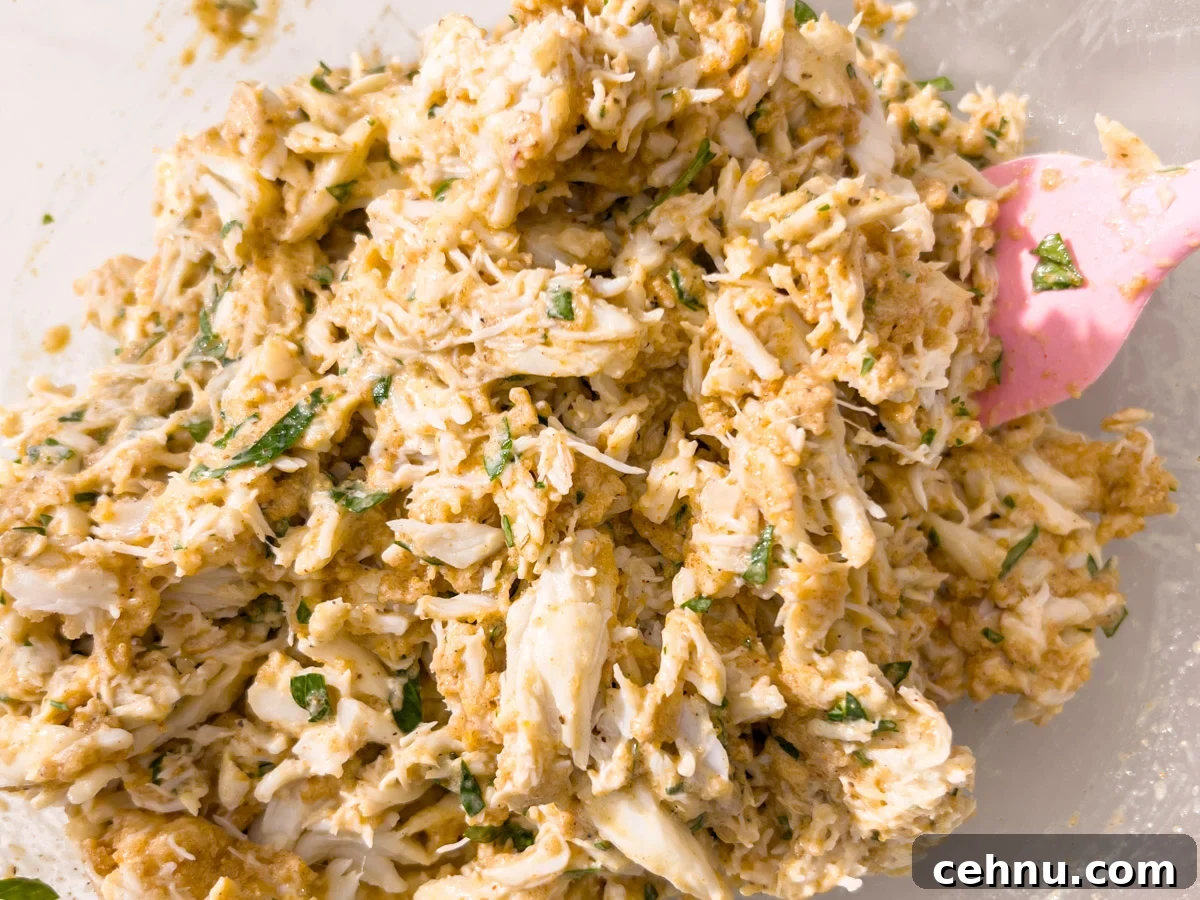 Crispy Gluten Free Crab Cakes Recipe with Lemon Dill Sauce 5 Fully mixed gluten-free crab cake filling with parsley and crab, being folded together with a pink spatula in a glass bowl.
