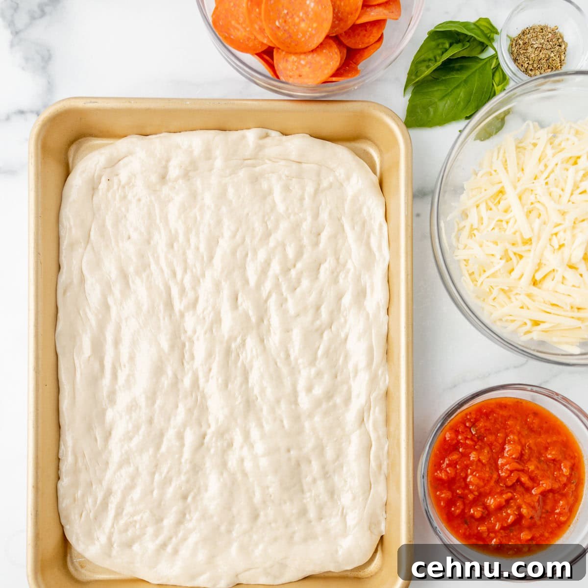 A rectangular sheet of pizza dough in a pan is surrounded by bowls of pepperoni slices, shredded cheese, tomato sauce, dried herbs, and fresh basil.