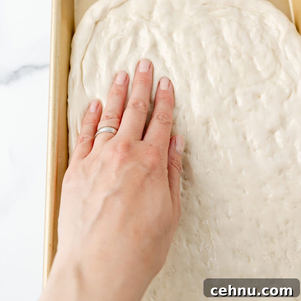 A hand pressing into risen bread dough in a rectangular pan on a white surface.