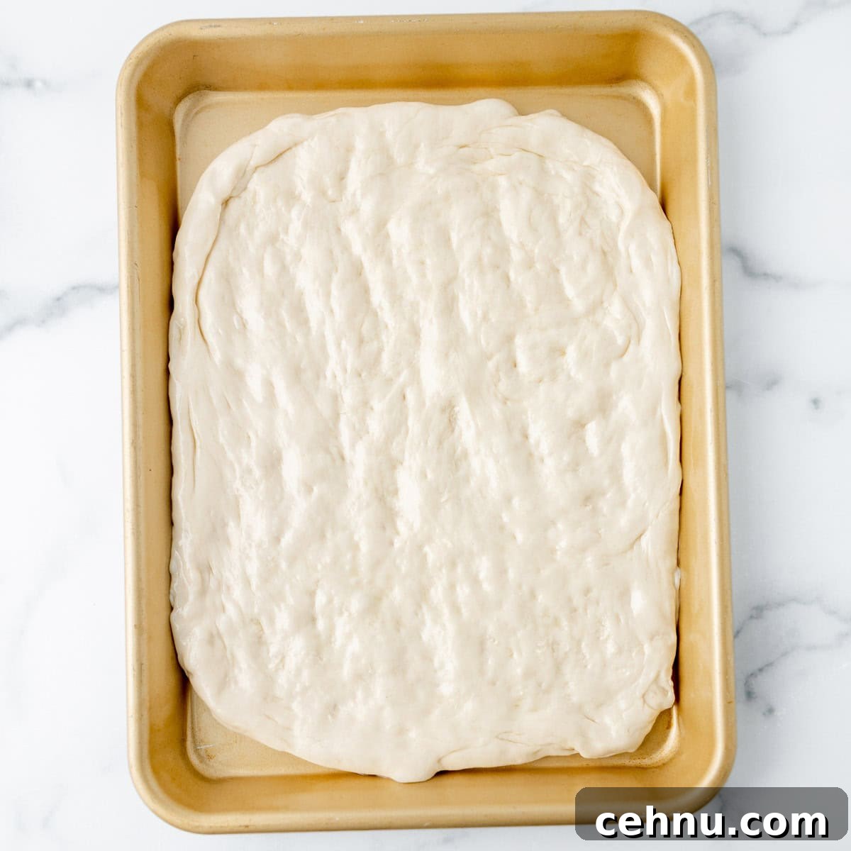Unbaked rectangular pizza dough spread evenly in a gold-colored baking sheet on a white marble surface.