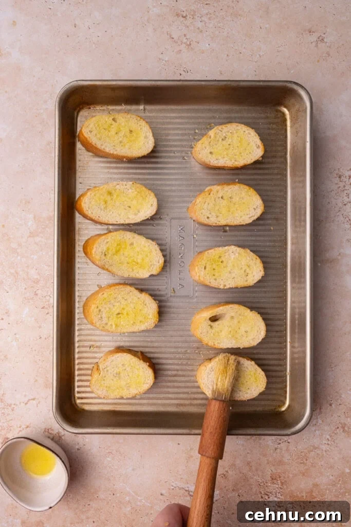 Sliced French baguette brushed with olive oil lined up on a baking sheet to toast in the oven.