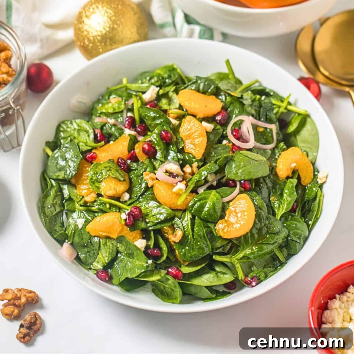 Salad made with baby spinach, mandarin oranges, pomegranate arils, candied walnuts, crumbled feta, shallots, and champagne vinaigrette in white bowl next to candied nuts and shallots.